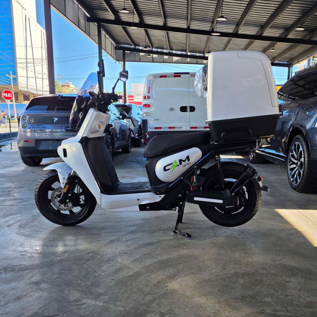 CAM 02 EV blanca: perfil lateral izquierdo completo con asiento negro, parabrisas y box trasero blanco en el dealer. Caja plástica blanca cerrada opcional: US$200 adicionales. Compatible con CAM 01 EV y CAM 02 EV; no incluida en el precio base de la moto. Parabrisas incluido con la CAM 02 EV.