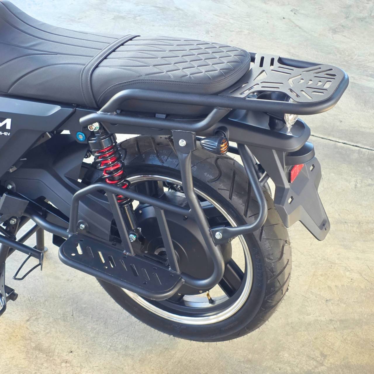 CAM 125 EV 2026 - Amarillo - Detalle de asiento, parrilla y rueda trasera. Corresponde a la misma unidad amarilla. El encuadre solo muestra componentes negros y el resorte rojo, no la carroceria amarilla.